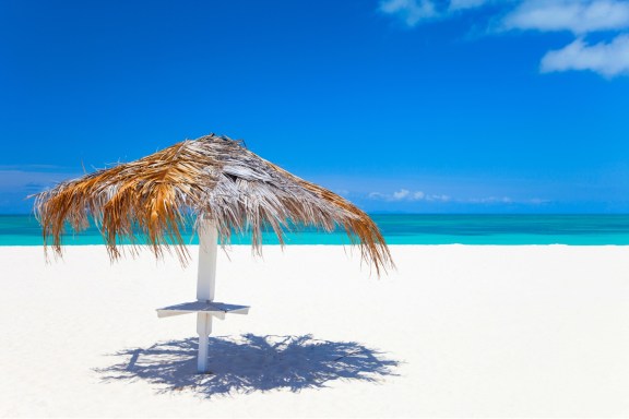 Snow white beach on Anegada Island, with a palm leaf umbrella overlooking the bright turquoise water and clear blue sky.