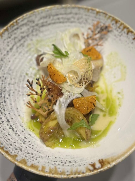 Amber's roasted baby cabbage, caramelized orange segments, and delicate fennel ribbons, complemented by vibrant herb oil and a light, creamy sauce. Garnished with edible coral tuile and fresh herbs.